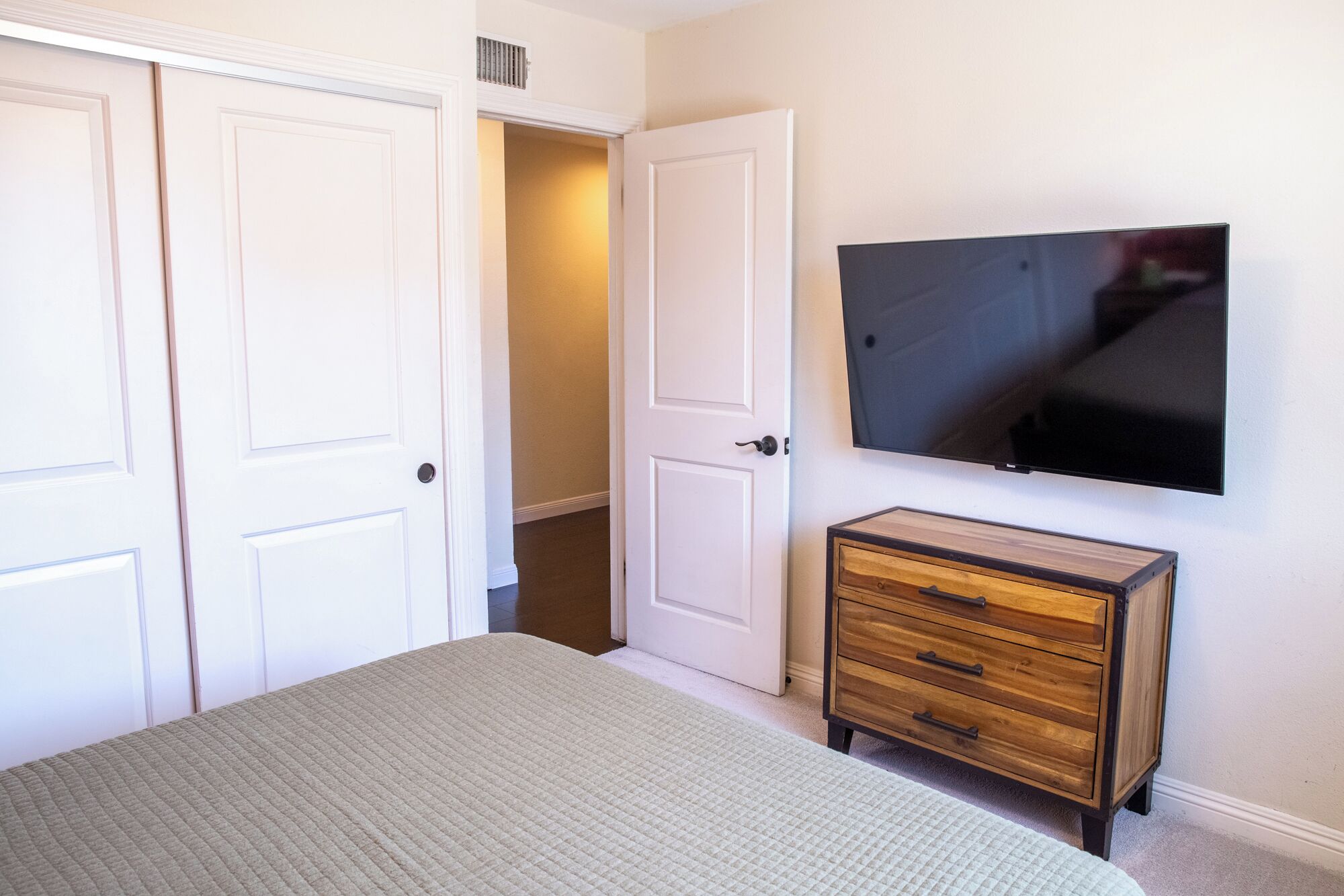 A bedroom with a bed, a dresser, a TV mounted on the wall, and a closet with two doors.