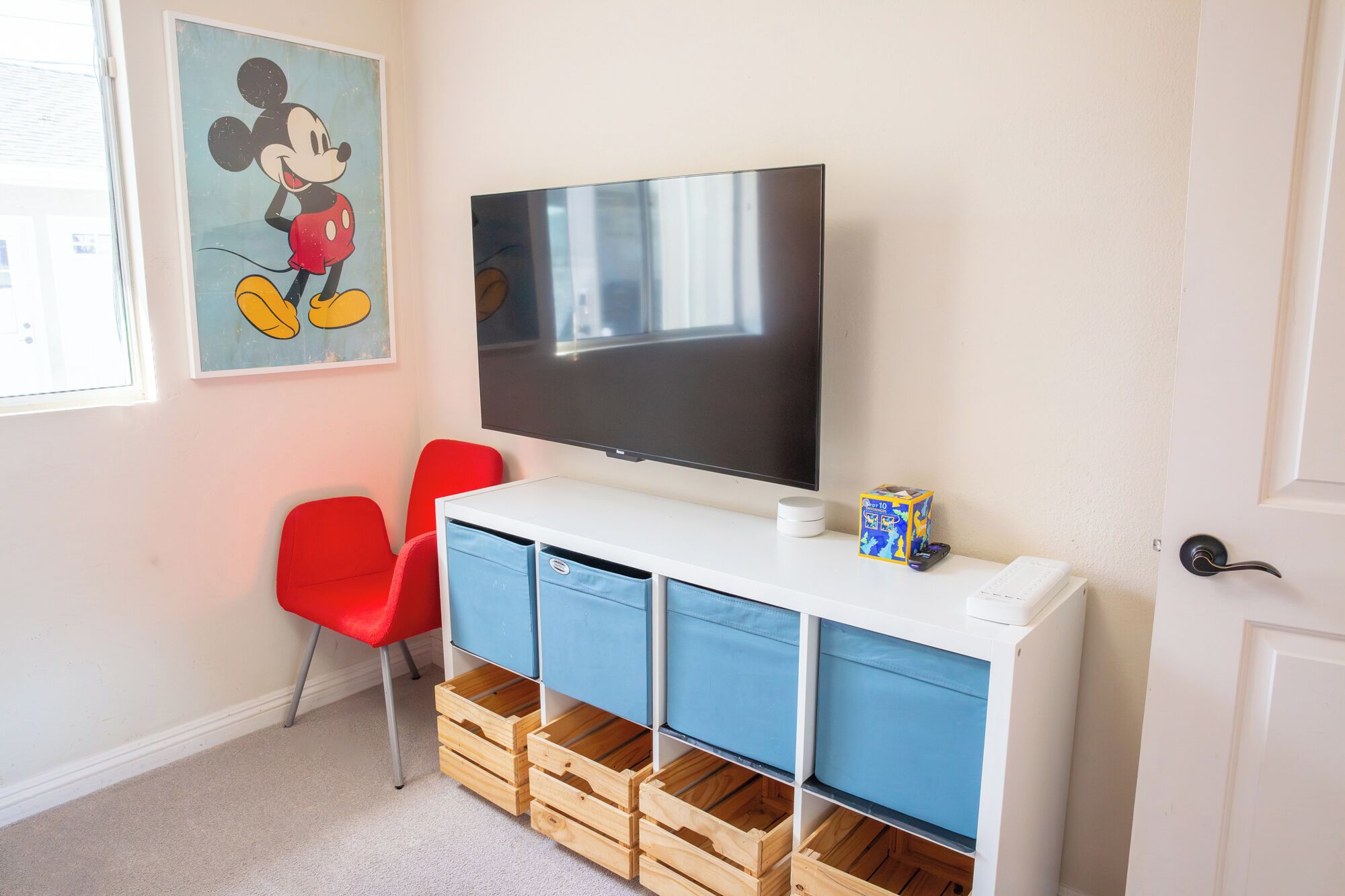 Una habitación con una televisión, sillas rojas, unidades de almacenamiento azules y un cuadro de Mickey Mouse.