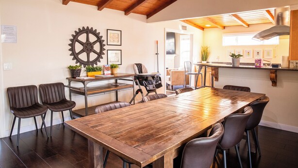 Un espace repas avec une table et des chaises en bois, un plafond haut avec des poutres apparentes, et une cuisine visible à travers une porte ouverte.