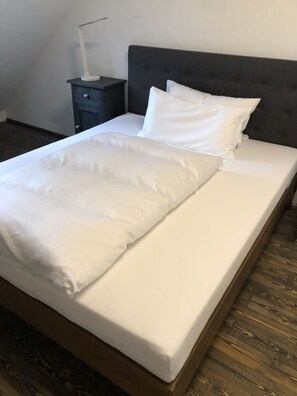 A neatly made bed with white linens in a room with wooden flooring and a dark headboard.