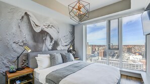 A modern bedroom with a large bed, a city view, a geometric light fixture, and a textured wall mural.