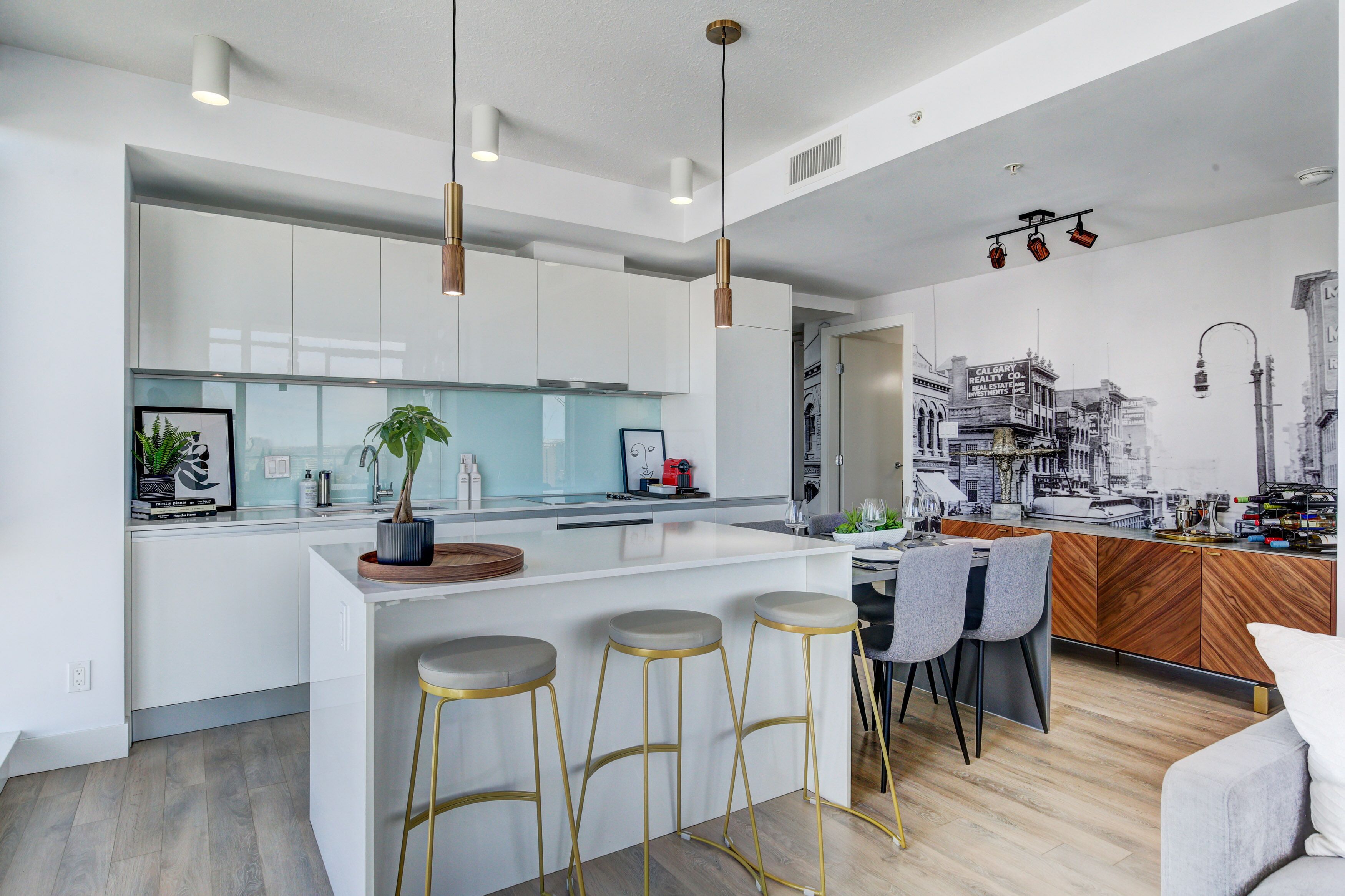 A modern kitchen with white cabinets, a central island with stools, pendant lighting, and a cityscape mural.