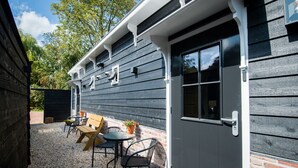 A building with a dark wooden exterior, a white door, and a small patio area with a table and chairs. - Cosy chalet in the heart of Monnickendam with terrace (Monnickendam)