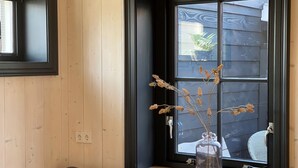A room with a wooden wall, a window with a view of a house, and a vase with dried flowers on a windowsill. - Cosy chalet in the heart of Monnickendam with terrace (Monnickendam)