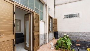 Un edificio residencial con puertas correderas abiertas que dan a una sala de estar, un pequeño jardín con plantas y un patio con baldosas.