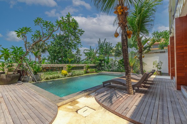 A wooden deck with a pool, surrounded by tropical plants and palm trees.