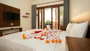 A hotel room with a bed, a TV, a window with curtains, and a view of greenery outside. - Sedana Cottage (Pererenan)