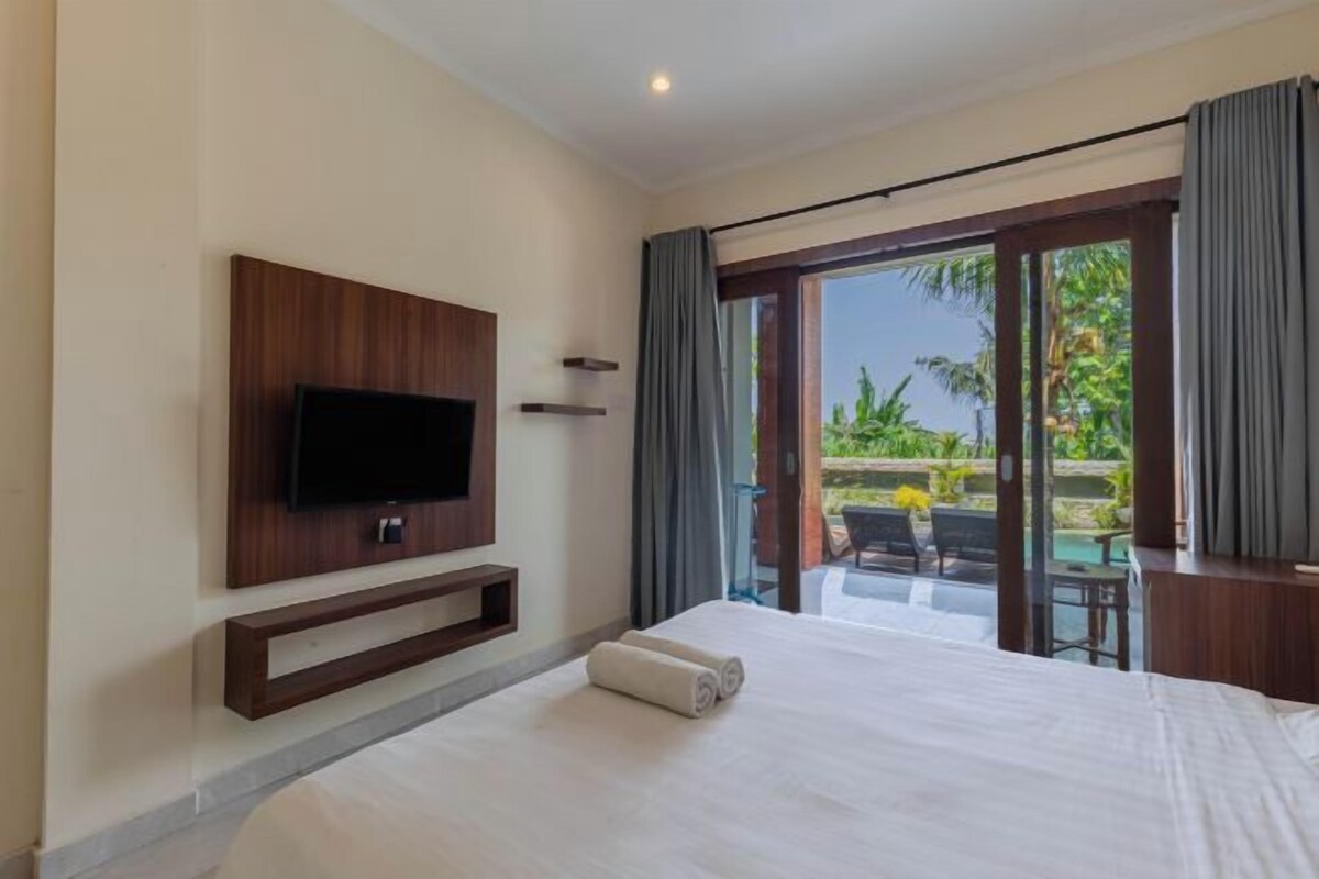 A hotel room with a bed, a flat-screen TV mounted on the wall, and a sliding glass door leading to an outdoor area with a pool and chairs.