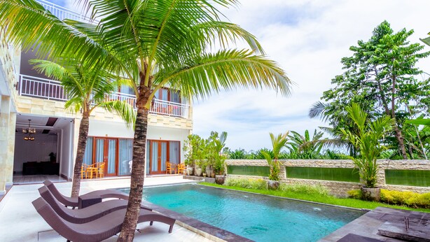 A swimming pool with lounge chairs, palm trees, and a two-story building with balconies. - Sedana Cottage (Pererenan)