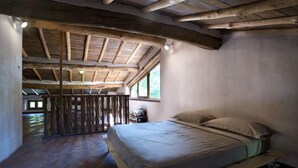 Una habitación rústica con techo de madera, una cama con colchón y almohadas, una mesita y una ventana que proporciona luz natural.