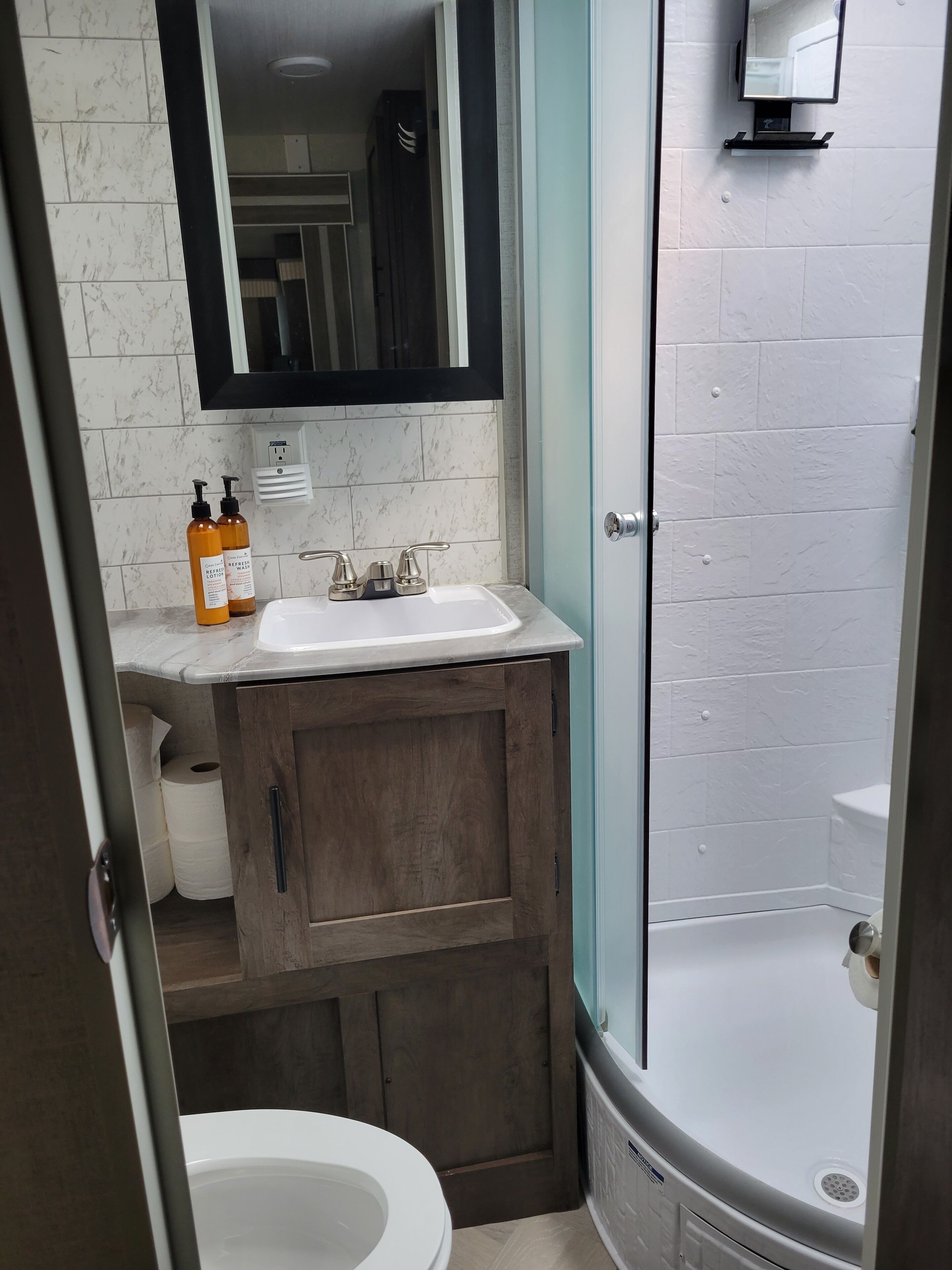 Ein Badezimmer mit Toilette, einem Waschbecken mit zwei Lotionflaschen, einem Spiegel über dem Waschbecken und einer Duschkabine mit einer weißen Badewanne.