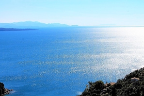 Une vue côtière sur une mer bleue et limpide, avec des montagnes lointaines et un petit village dont les bâtiments sont construits sur une colline.