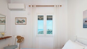 A bedroom with a bed, a chair, a window with curtains, and a wall-mounted air conditioner. - Two bedroom apartment with breathtaking views (Corfu)