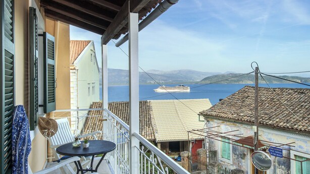 A balcony with a table and chairs, overlooking a body of water with a cruise ship in the distance and buildings with a Mediterranean architectural style. - Two bedroom apartment with breathtaking views (Corfu)