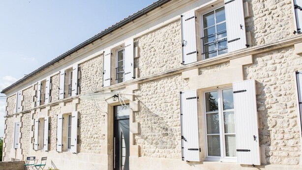 Un edificio de piedra con varias ventanas y contraventanas, una puerta y asientos exteriores.