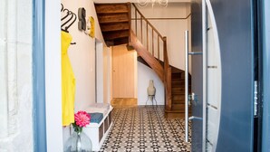 Un pasillo con suelo estampado, una escalera de madera, una araña y un banco con un jarrón de flores.