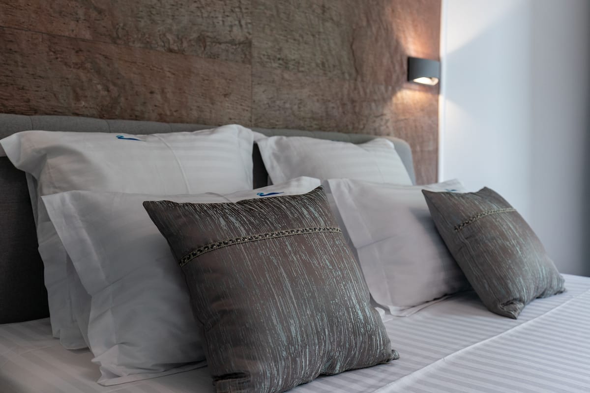 A neatly made bed with white and gray pillows, a wooden headboard, and a wall-mounted light fixture.