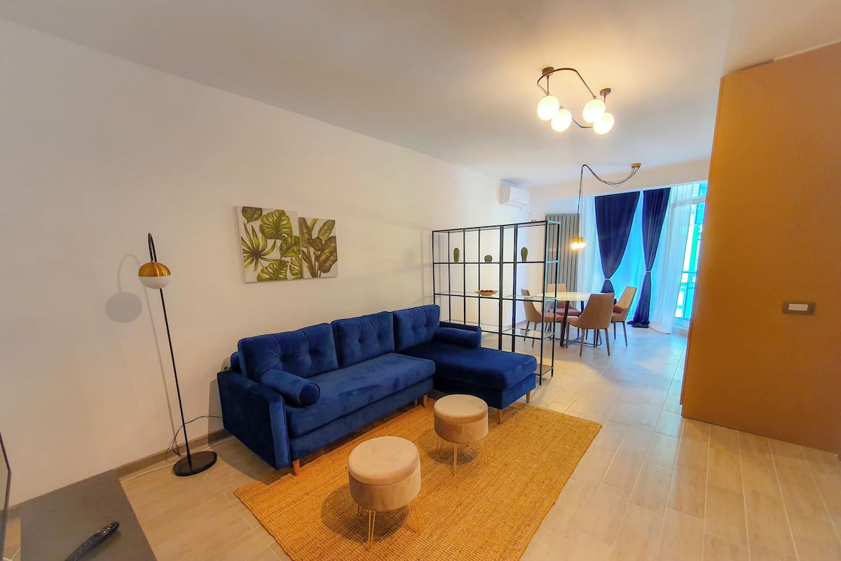 A modern living room with a blue sectional sofa, a small round ottoman, and a floor lamp. There is a dining area with a table and chairs in the background.