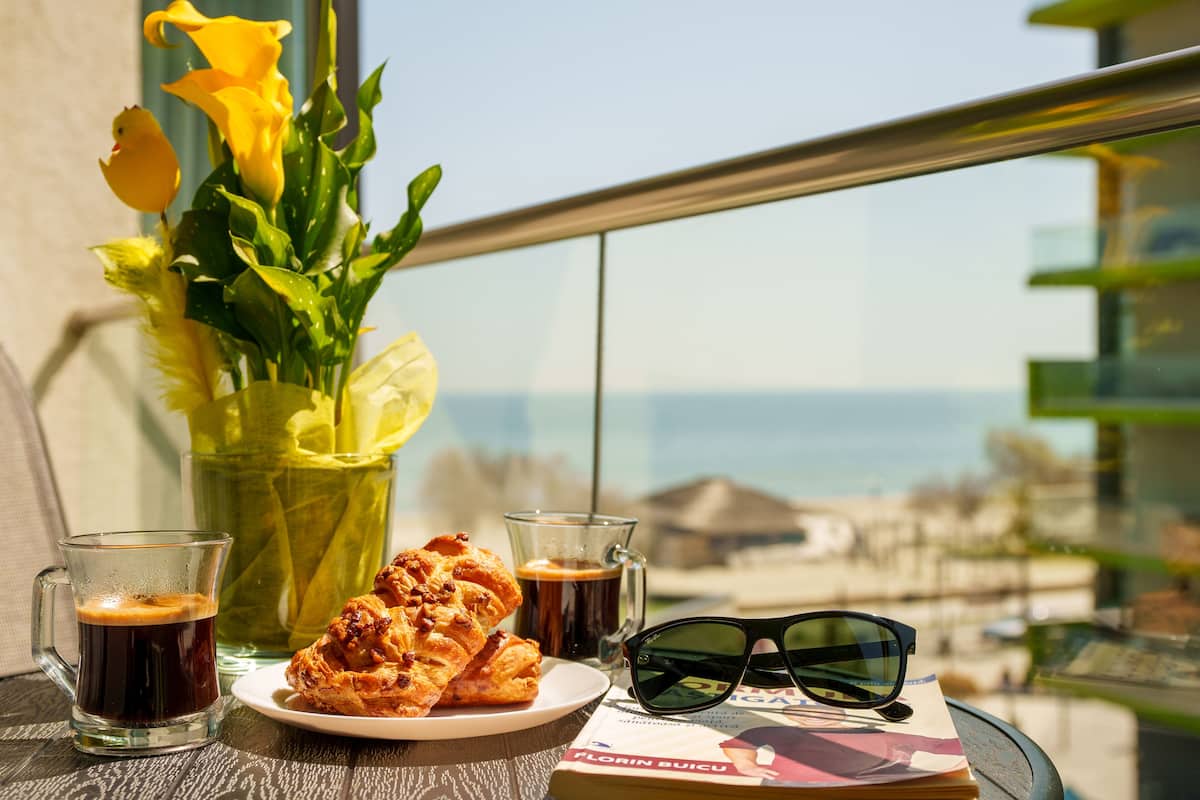 A table with a plate of pastries, two glasses of coffee, a vase with yellow flowers, and a pair of sunglasses on a magazine.