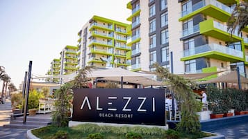A modern building with balconies, a sign reading "ALEZZI BEACH RESORT," and outdoor seating.