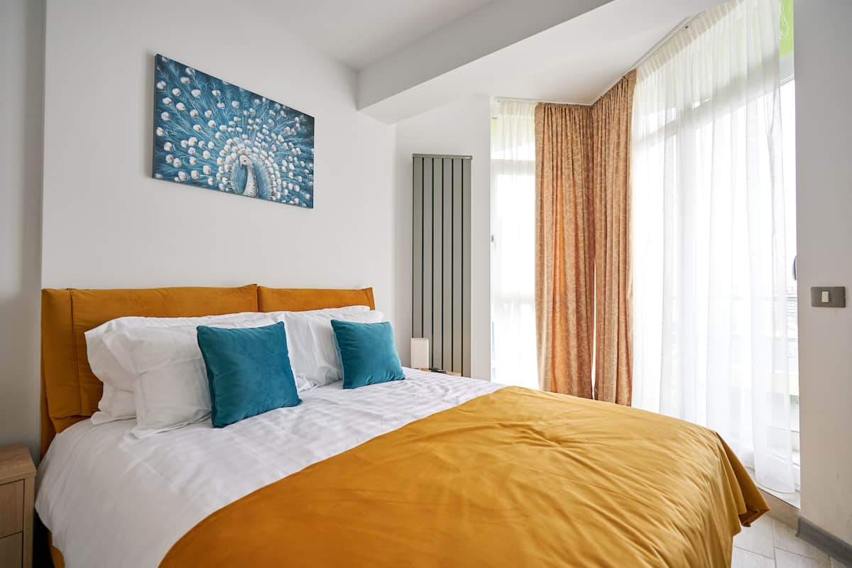A hotel room with a large bed, a peacock painting on the wall, a radiator, and a window with curtains.