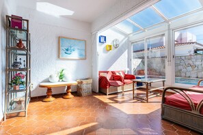 A sunlit room with a glass door leading to a pool, a wicker sofa, a small table, and a decorative shelf.