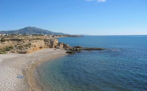 Une plage côtière aux eaux cristallines, avec un rivage rocheux et une montagne qui se profile à l’horizon.