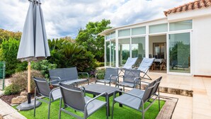 Una terrazza esterna con tavolo e sedie, un ombrellone grande e una stanza con vetrate che si affaccia sul giardino.