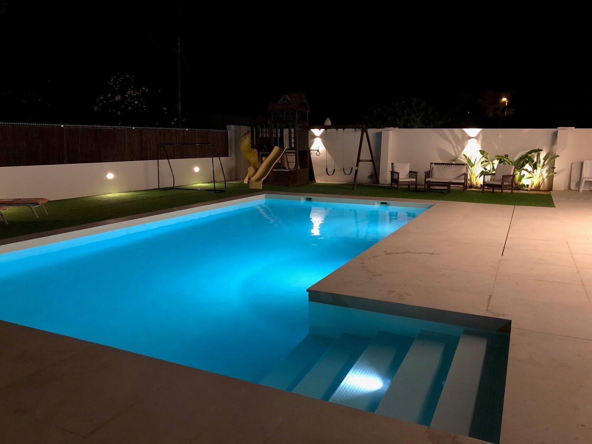 A swimming pool with a slide, surrounded by a white wall and a small garden area with chairs and plants.