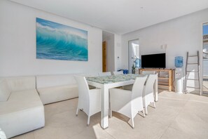 A modern living room with a white sofa, a dining table with white chairs, a large TV, and a water dispenser.