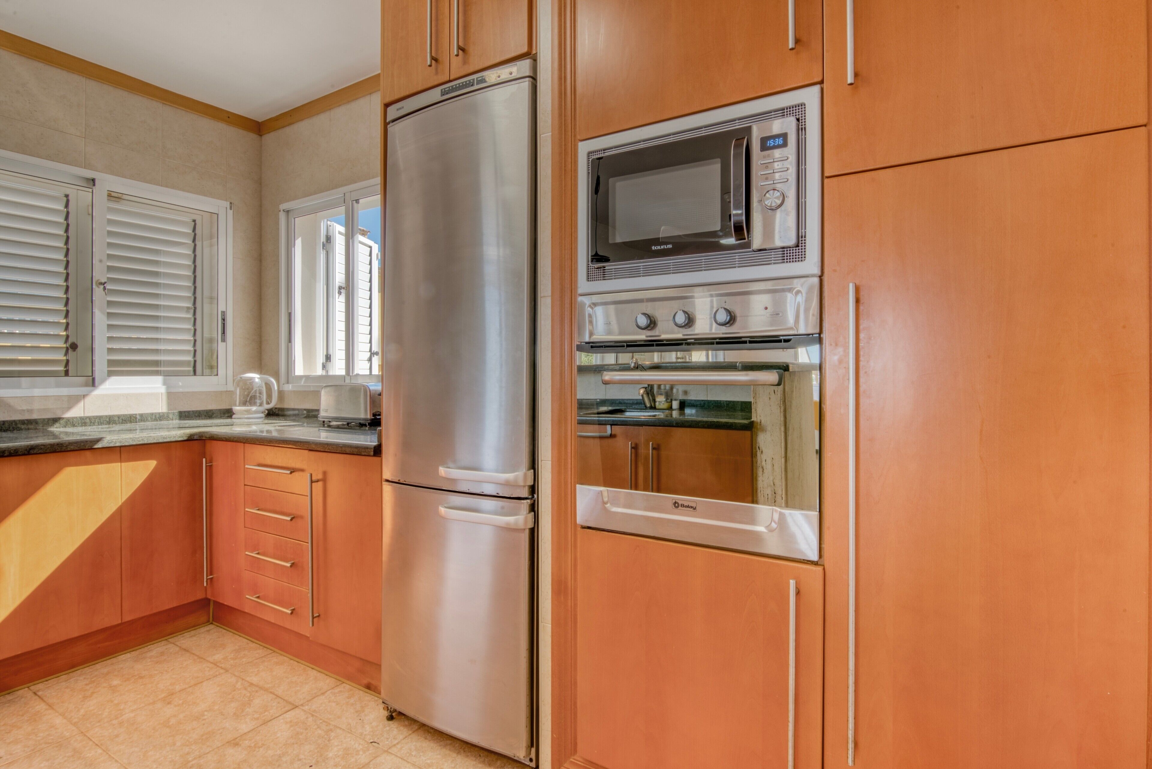 Una cocina con electrodomésticos de acero inoxidable, armarios de madera y un ventanal con persianas.