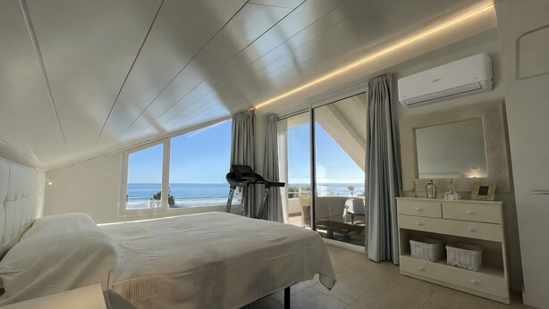 Une chambre à coucher avec un lit, un tapis de course, une grande fenêtre donnant sur la mer, une commode blanche et un ventilateur de plafond.