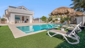 Une maison moderne dotée d’une piscine, d’un espace salon extérieur et d’une structure surmontée d’un toit de chaume.
