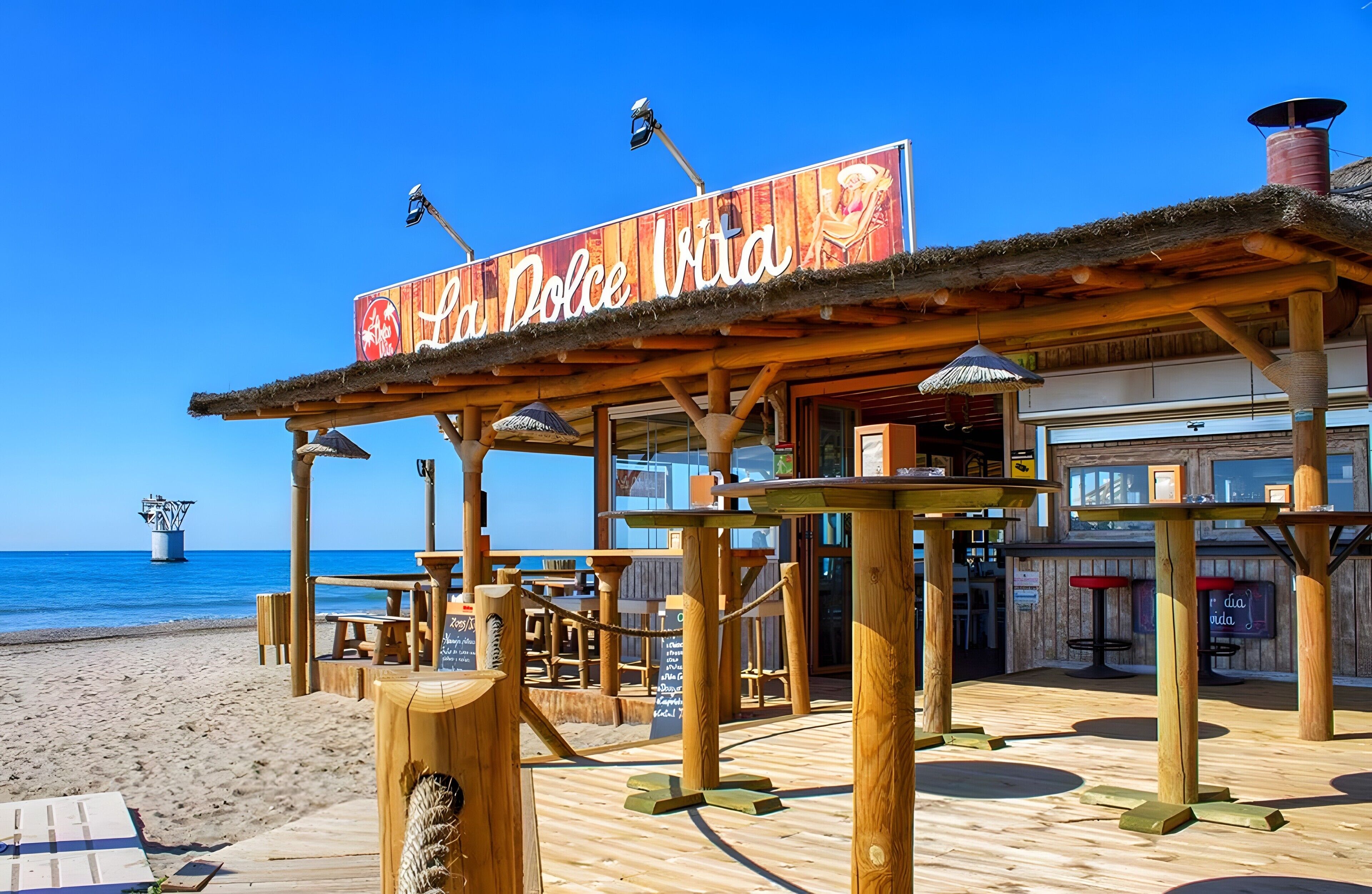 Ein hölzernes Café am Strand, mit einem Schild mit der Aufschrift 'La Dolce Vita' und Blick aufs Meer.