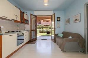 A kitchen with white cabinets, a stove, and an oven. There is a doorway leading to a balcony with a view of a garden and a building. A brown sofa is in the living area.