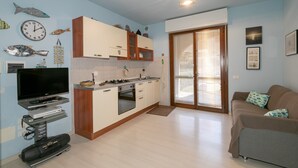 A modern kitchen with a TV, oven, and cabinets, a living area with a sofa, and a sliding glass door to the outside. - Studio Residence Valle Fiorita with Shared Pool & Wi-Fi in Riotorto (Piombino)