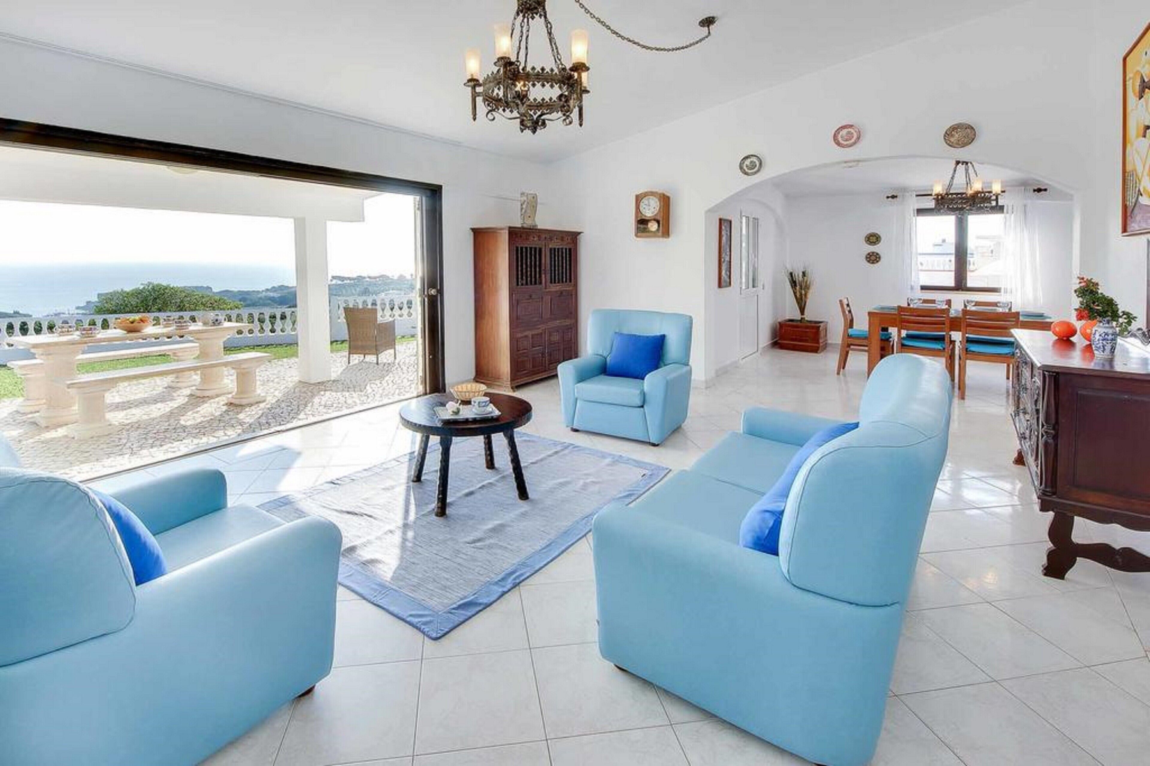 A spacious living area with a large window, a chandelier, and a dining area with wooden furniture.