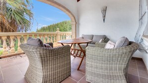 Une terrasse couverte, aménagée avec des meubles en osier, une table et offrant une vue sur les arbres et le ciel.