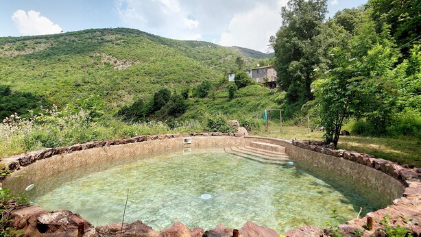 Ein klares Schwimmbecken, umgeben von Steinmauern, eingebettet in eine üppige grüne Landschaft mit Hügeln im Hintergrund.