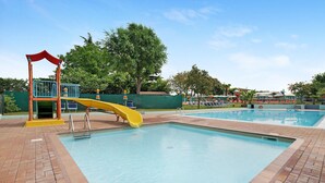 Une piscine avec un toboggan jaune, un poste de surveillance pour les sauveteurs et un espace de repos abrité.