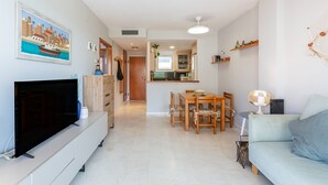 Uma sala de estar moderna com uma TV de tela plana, uma área de jantar com mesa e cadeiras, e uma cozinha ao fundo.