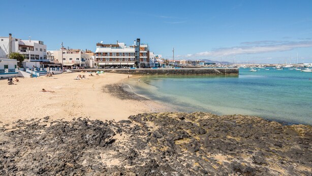 Un pueblo costero con una playa de arena, un litoral rocoso y edificios con balcones que dan al mar.
