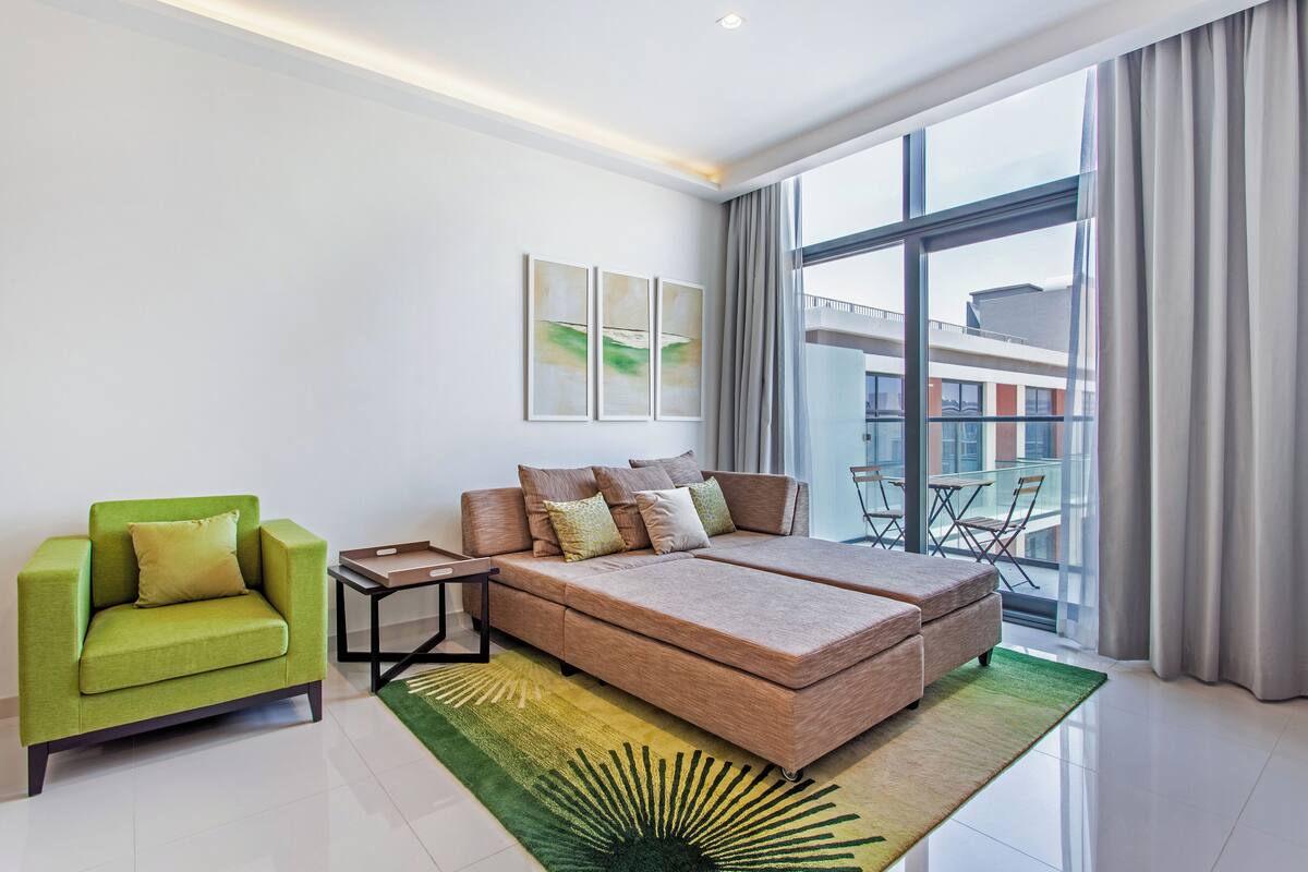 A modern hotel room with a large bed, a green armchair, and a view of the city through large windows.