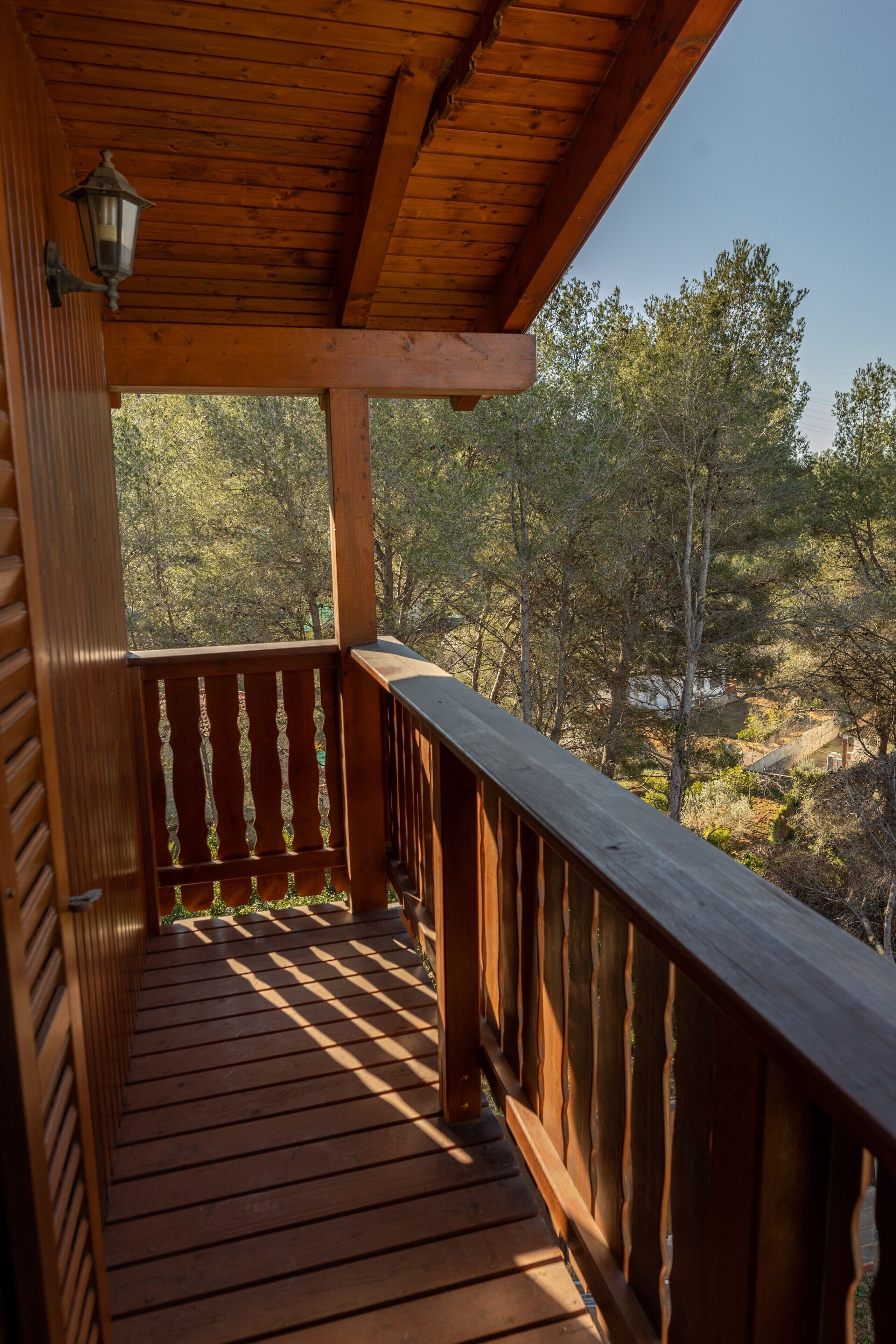 Un balcón de madera con barandilla, un farol y vistas a árboles y un paisaje.