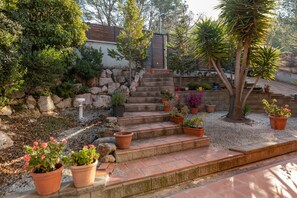 A garden with steps leading to a door, surrounded by various potted plants and a palm tree. - Chalet 'Casa de Madera' with Mountain View, Garden, A/C, Pool & Wi-Fi (Calafell)