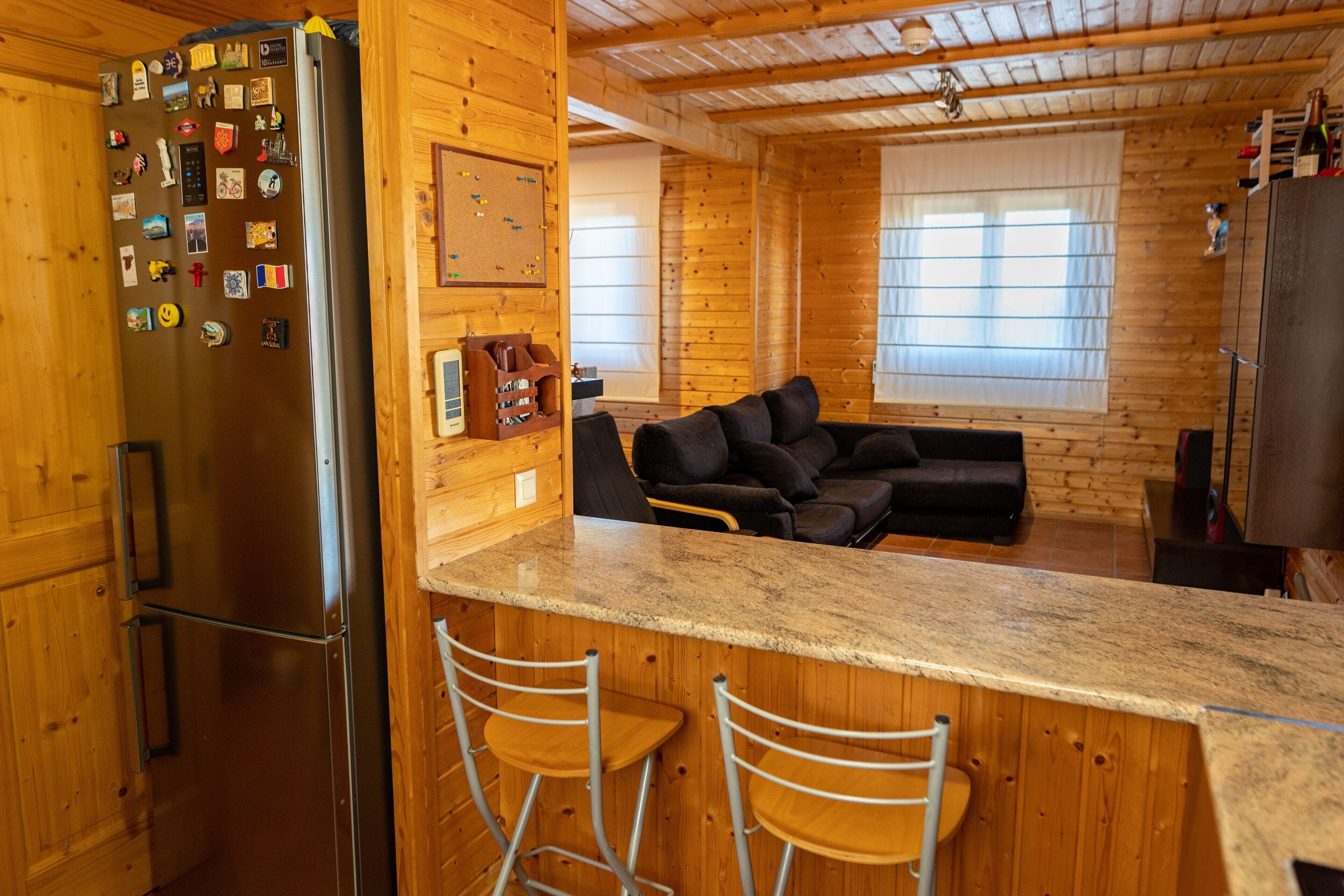 Een houten blokhut van binnen, met een keukenhoek, een bar met krukken, een zwarte bank en een prikbord aan de muur.
