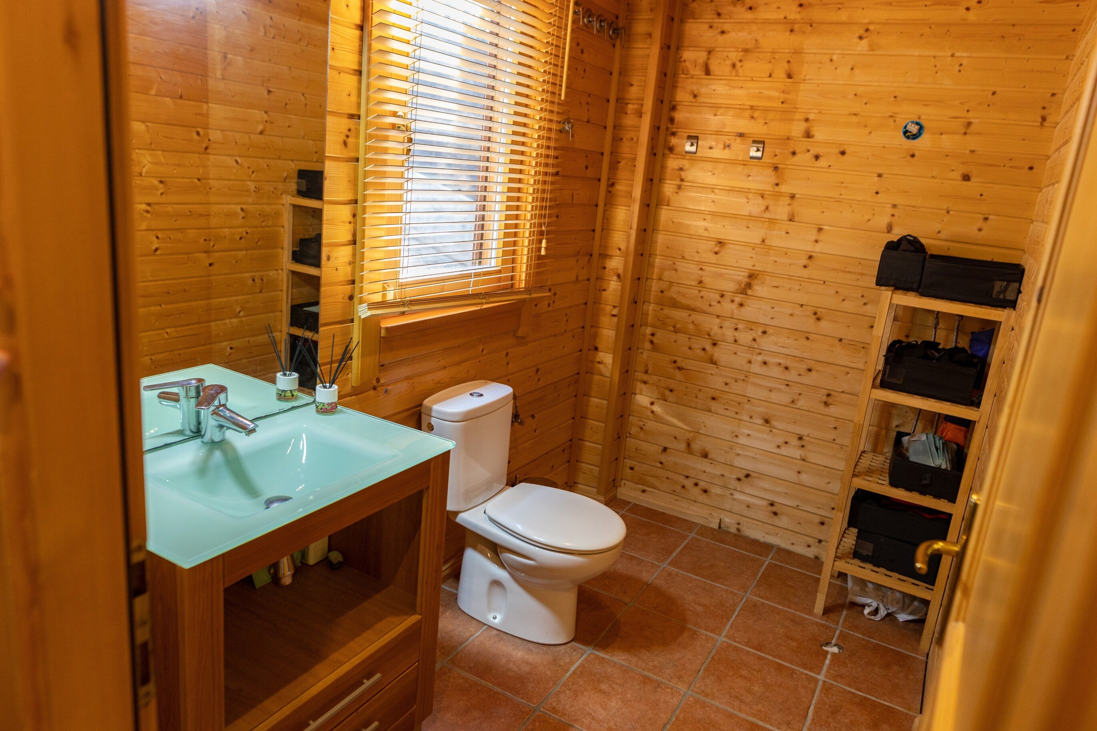 Een houten badkamer met een glazen wastafel, een wit toilet en een houten plank met opbergdozen.