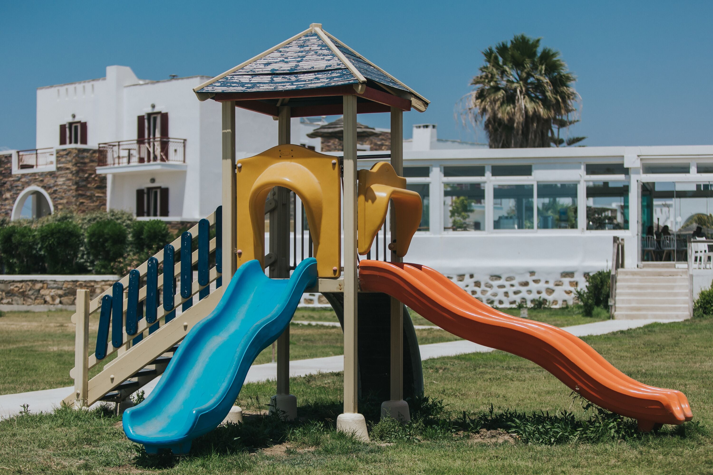 Un parque infantil con toboganes y un refugio, frente a un edificio de fachada de piedra y un palmera al fondo.