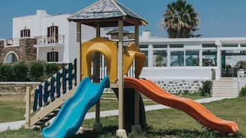 Un parque infantil con toboganes y un refugio, frente a un edificio de fachada de piedra y un palmera al fondo.