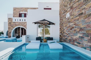 A modern pool area with a stone wall, a hanging chair, and a lounge chair. - Aegean Palace (Naxos)